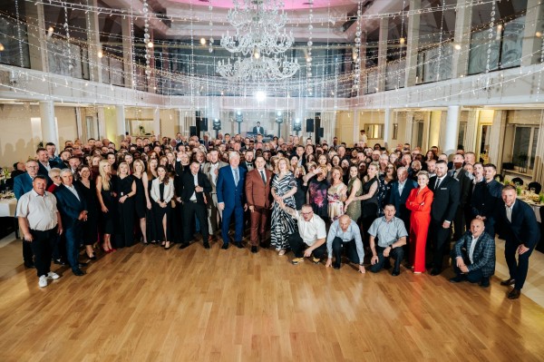 Panoramiczne zdjęcie dużej grupy osób – prawdopodobnie pracowników firmy – zebranych w sali bankietowej na uroczystej kolacji lub gali. Uczestnicy ubrani są w eleganckie stroje wieczorowe i pozują do wspólnej fotografii na drewnianym parkiecie. Sala jest bogato udekorowana girlandami światełek i dużym, kryształowym żyrandolem. Zdjęcie symbolizuje zintegrowany zespół i wydarzenie firmowe.