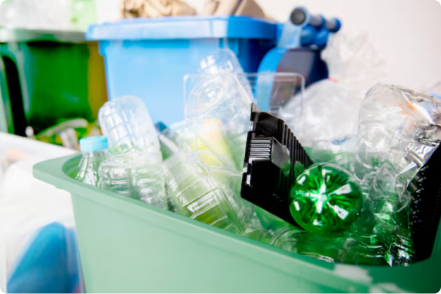 Zbliżenie na zielony pojemnik wypełniony po brzegi przezroczystymi butelkami PET, plastikowymi opakowaniami i tacami. W tle, rozmyte, widoczne są inne pojemniki do segregacji odpadów, w tym niebieski. Obraz przedstawia dużą ilość zebranego plastiku przeznaczonego do recyklingu.